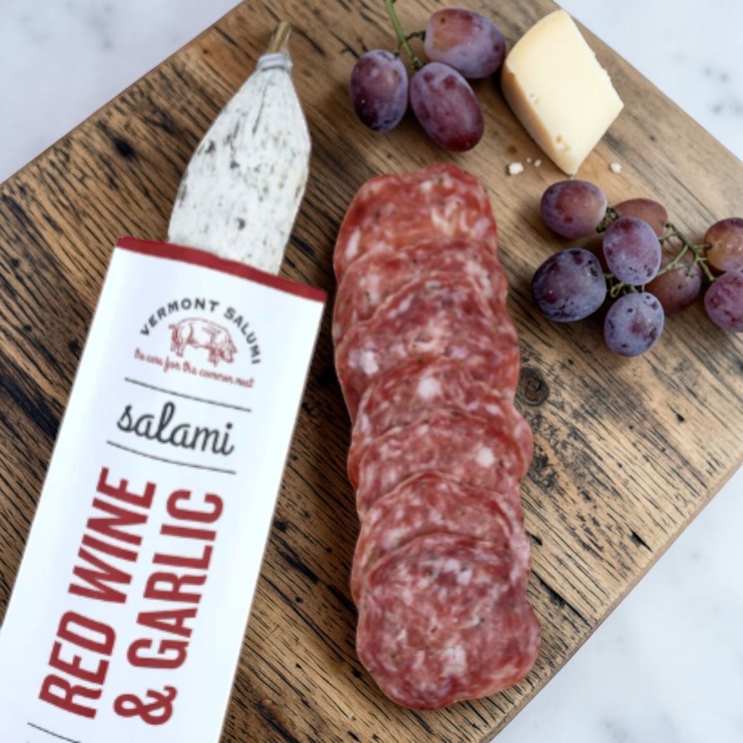 Stowe Public House & Bottle Shop Vermont Salumi Red Wine & Garlic salami, sliced and whole, with grapes and cheese on a wooden board.