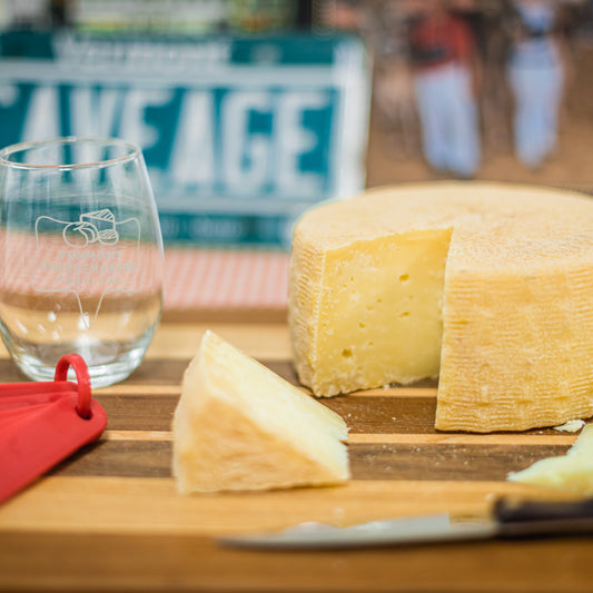 Stowe Public House & Bottle Shop Chin Clip raw milk cheese wheel, aged four months, with a natural rind and complex flavor. Served on a wooden board with a wine glass.